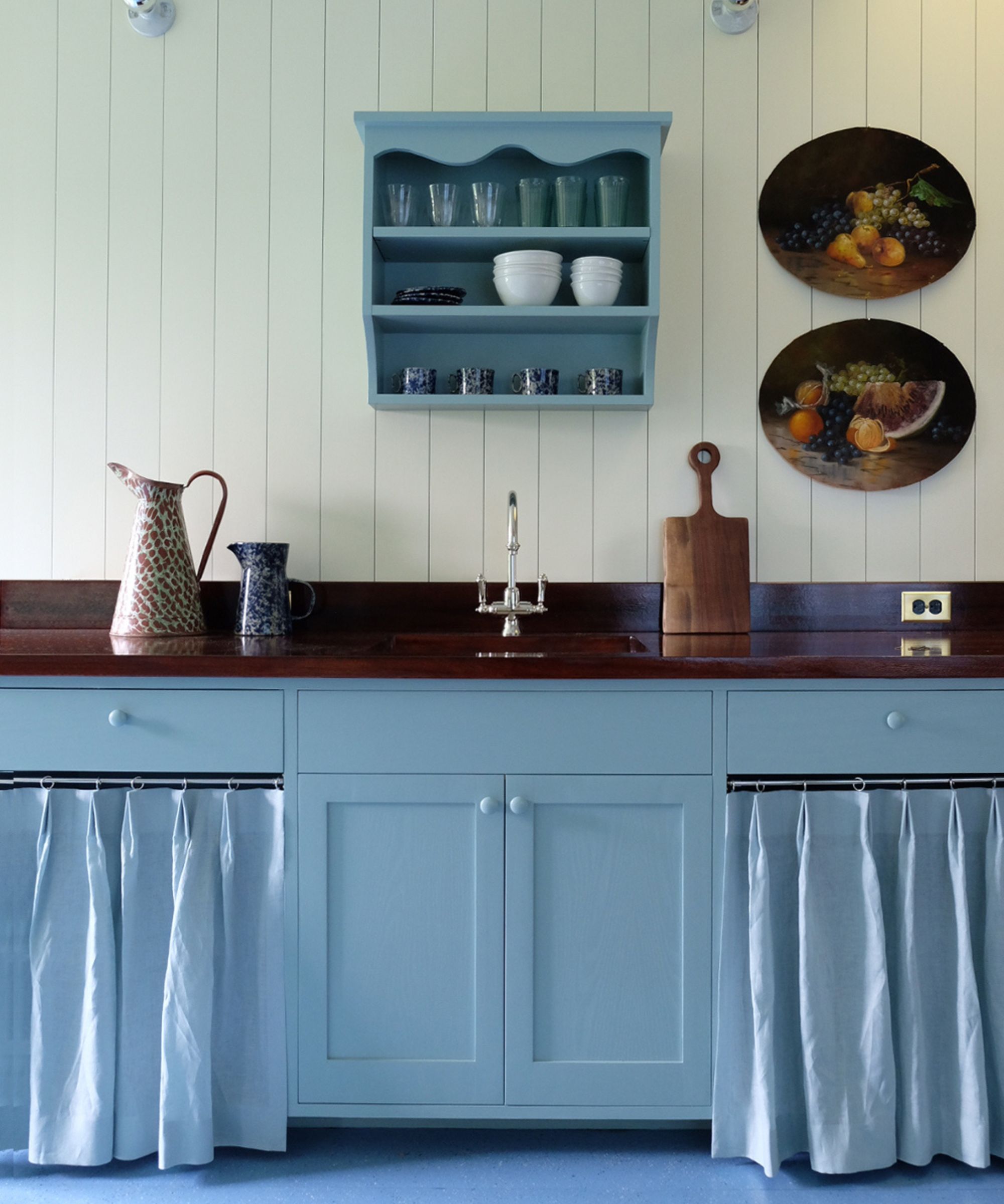 a New England cottage with paneled walls and a light blue kitchen with dark wooden counters, cabinet curtains and a blue painted floor