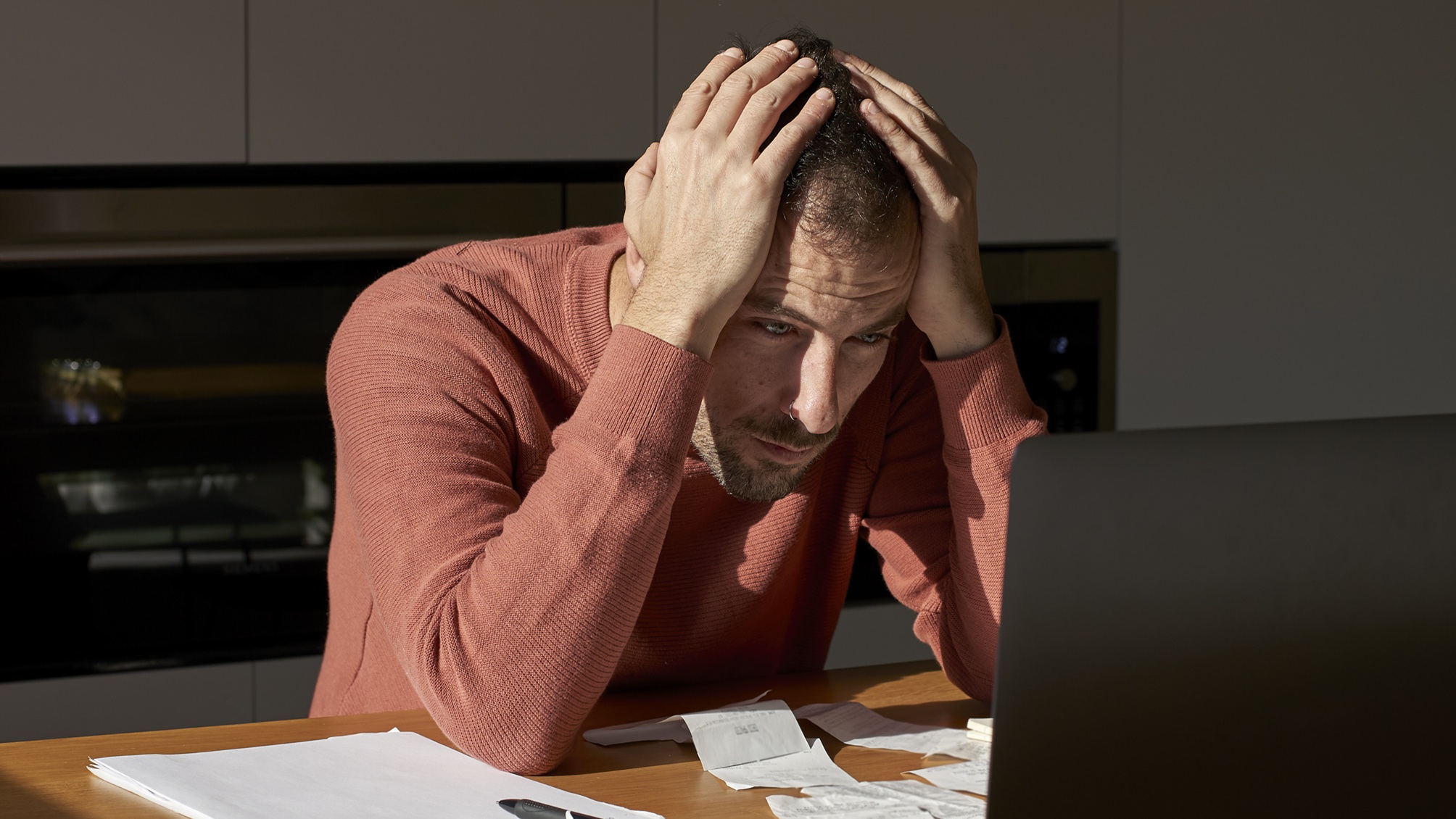 A stressed man holds his head in his hands as he looks at his laptop.