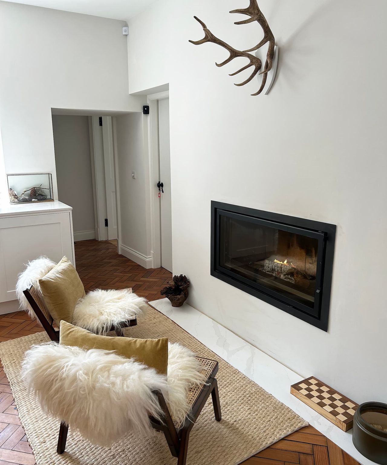 Two chairs facing an fireplace inbuilt into a white wall with a pair of deer antlers hanging above