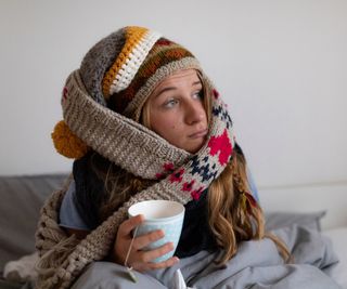 young woman wearing scarf and hat sat in bed drinking tea looking sad