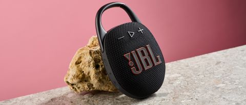 The JBL Clip 5 in black is pictured leaning against a sand colored rock against a pink background,