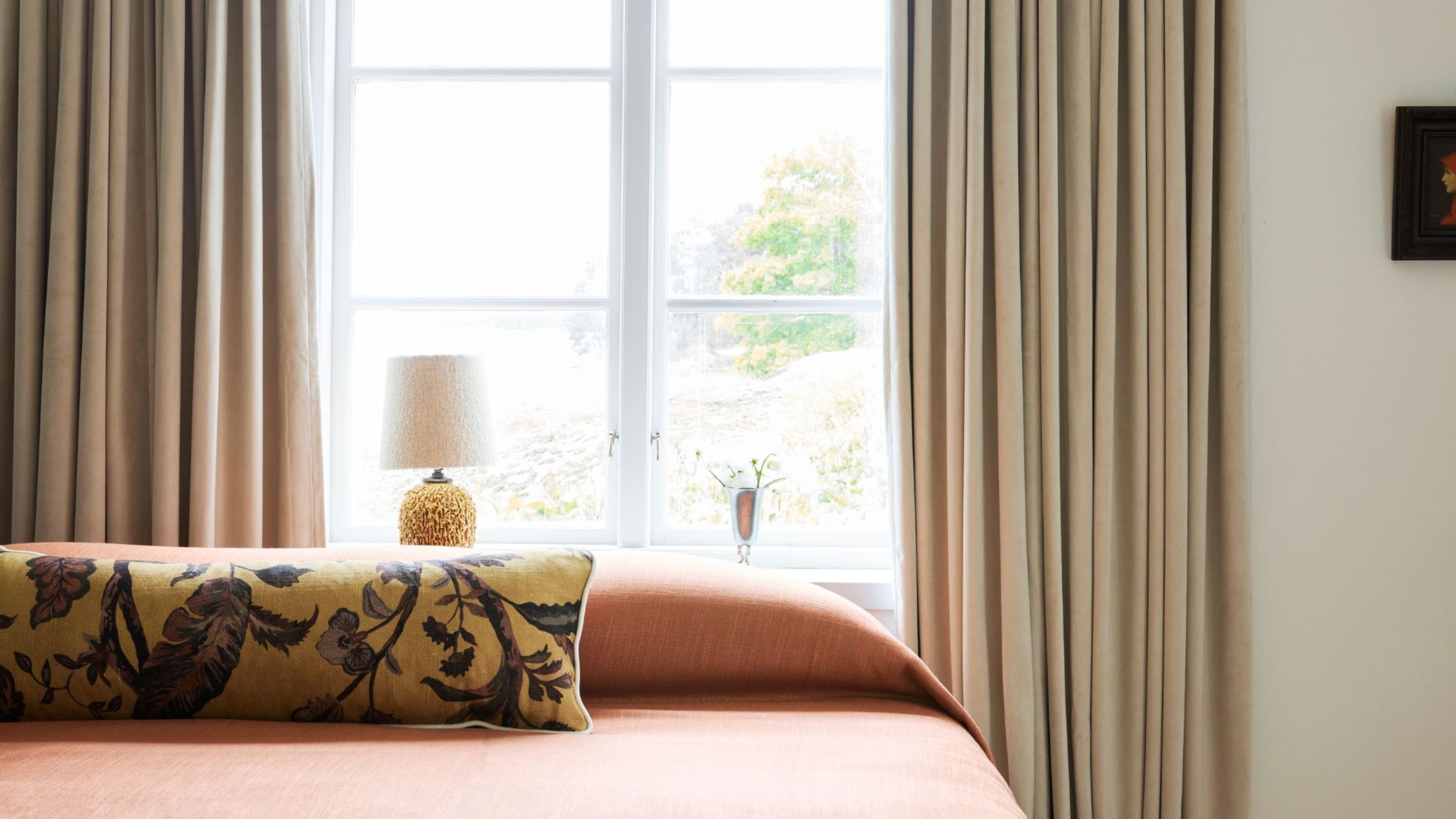 modern bedroom with bed dressed in orange bedcover and patterned bolster pillow, with window behind the bed dressed with draped curtains and a lamp on the windowsill 
