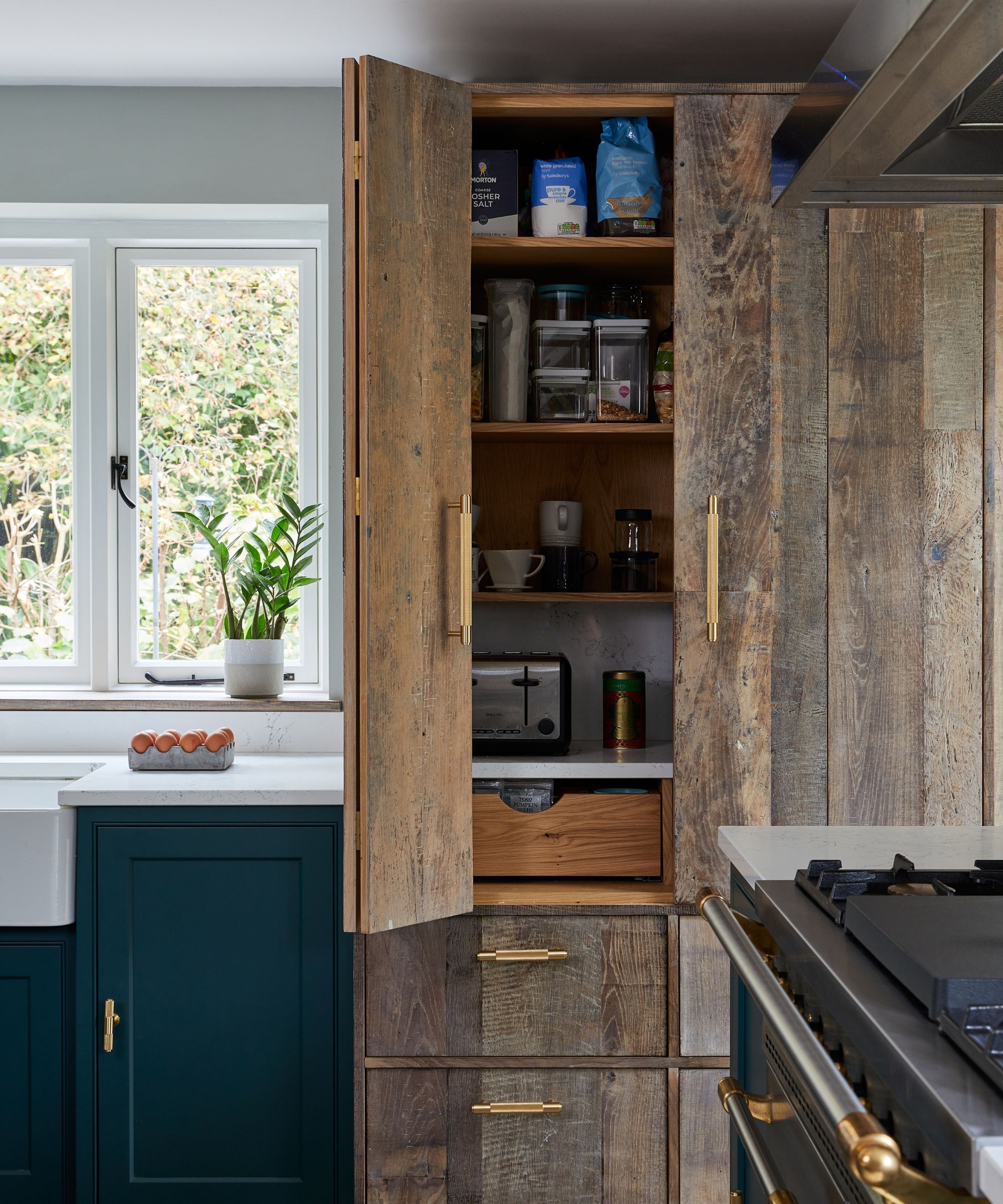 Rustic-modern pantry with weathered wood doors, brass handles, and oak shelving, blending natural textures with sleek kitchen cabinetry and soft daylight.