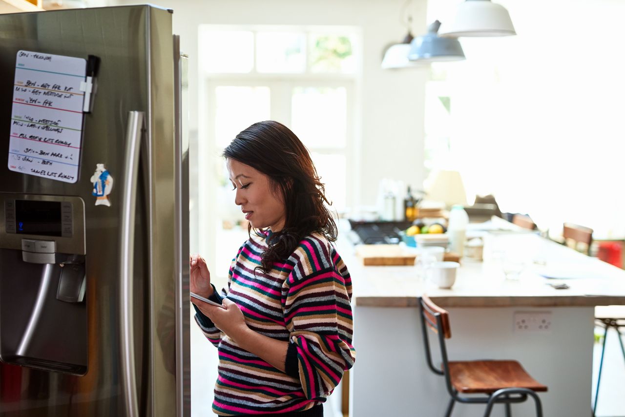 woman looking in fridge while holding phone - illustrating checking what&#039;s in the fridge while doing online food shop