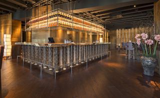 A square bar counter made from natural wood with bamboo shaped lights on the sides and dark wooden floors.