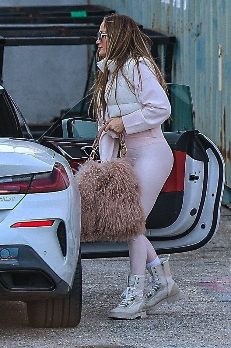 Jennifer lopez wears a white puffer vest, pale pink sweatshirt, pale pinks leggings, pink fur back, and white combat boots.