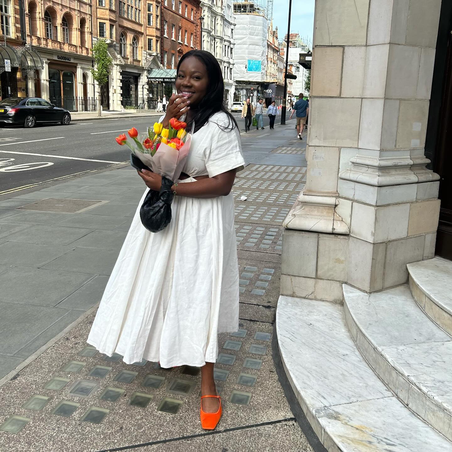 Iman wears short-sleeve white sundress and orange flats while holding a black handbag and flowers