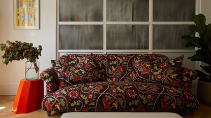 patterned sofa with dark floral print in front of fluted glass internal windows, with a red arnold circus stool next to the sofa with a vase and branch on it and an artwork hanging on the wall above