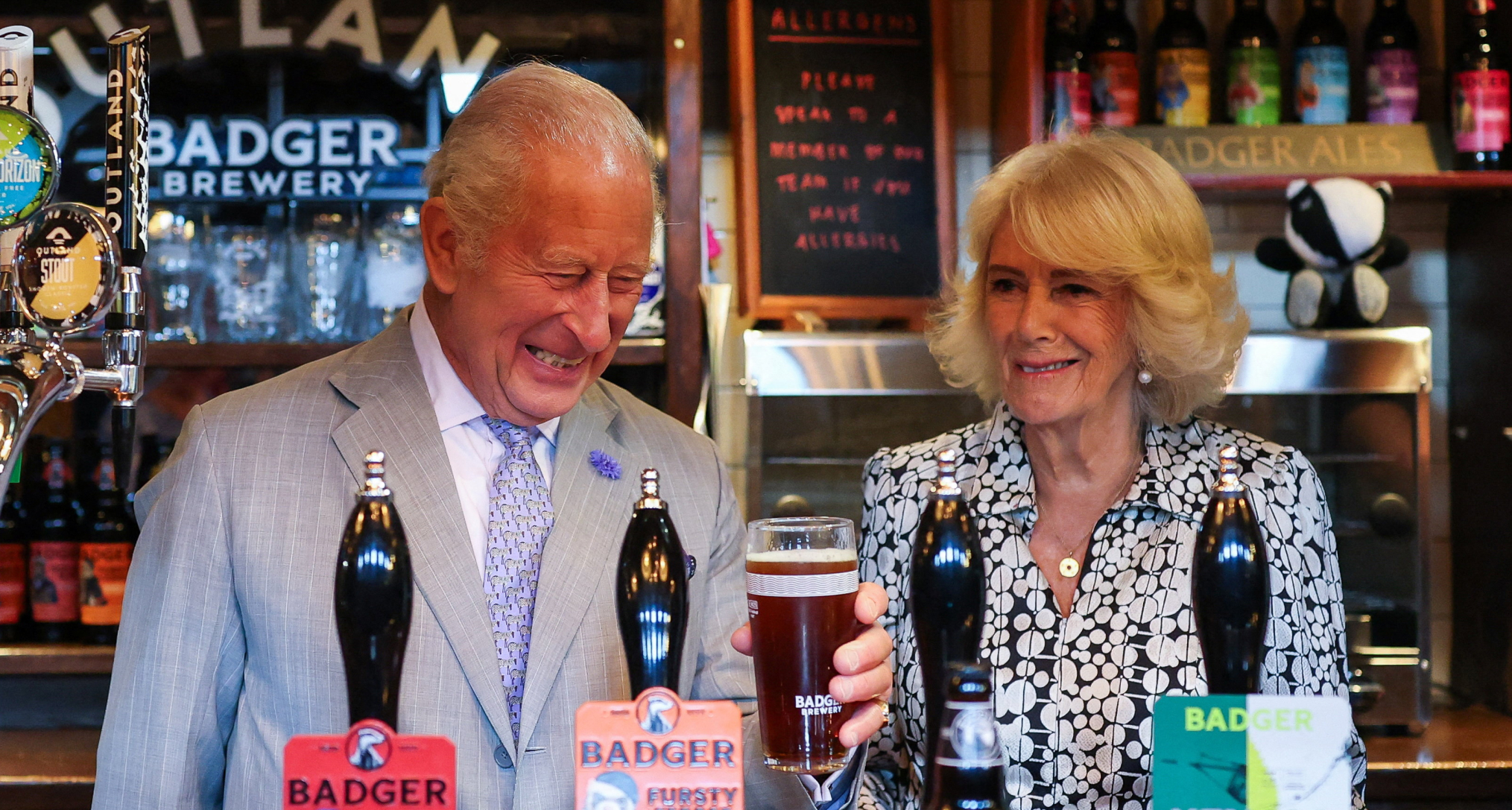 Queen Camilla and King Charles laughing as they pull pints at a pub
