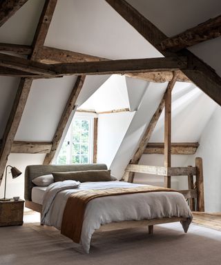 A bedroom in a loft conversion featuring exposed wooden beams and a pitched ceiling. The room includes a simple wooden-framed bed with white bedding, a rust-coloured throw, and a grey upholstered headboard.