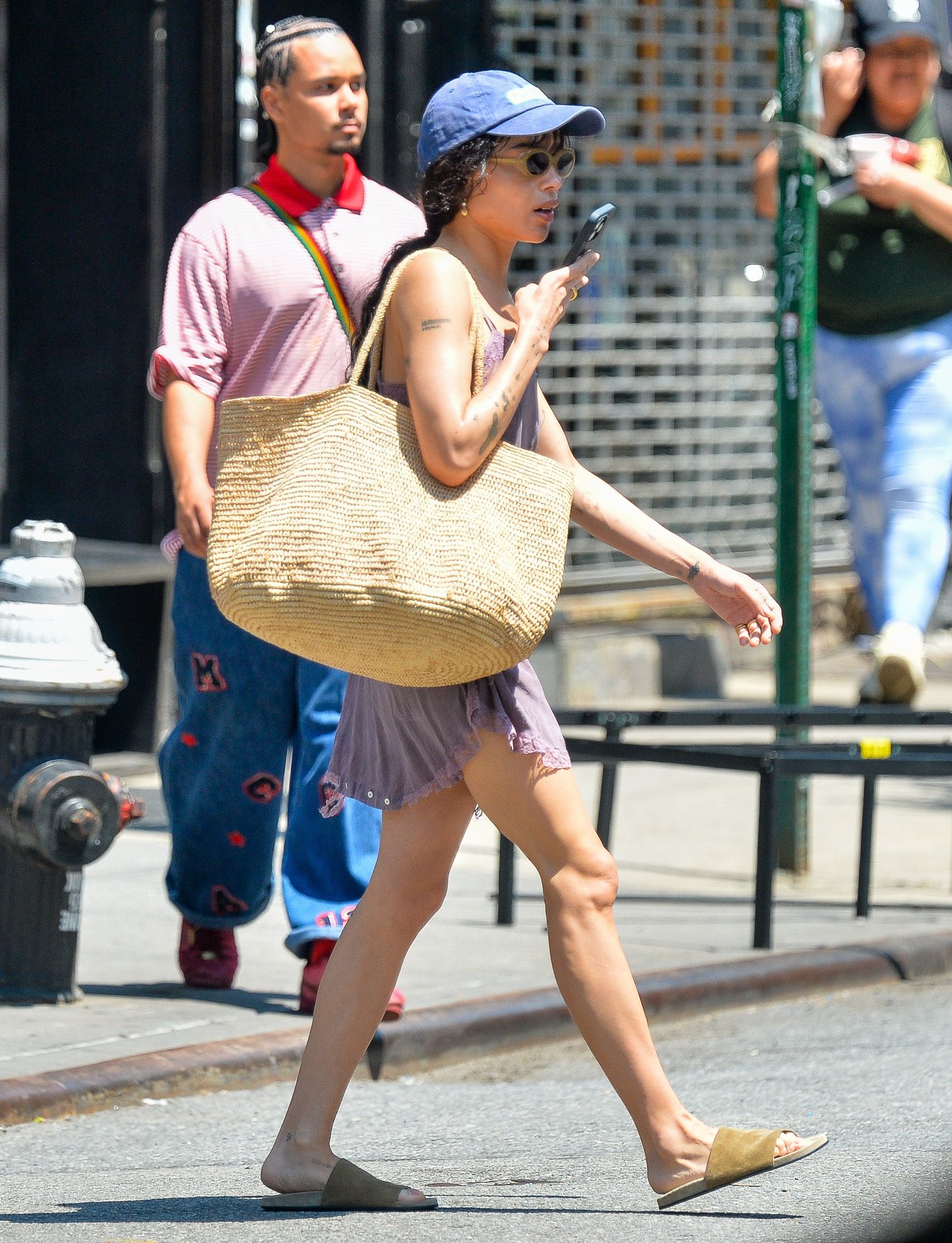 Zoe wears suede flats, a raffia bag, dress, and blue baseball cap.