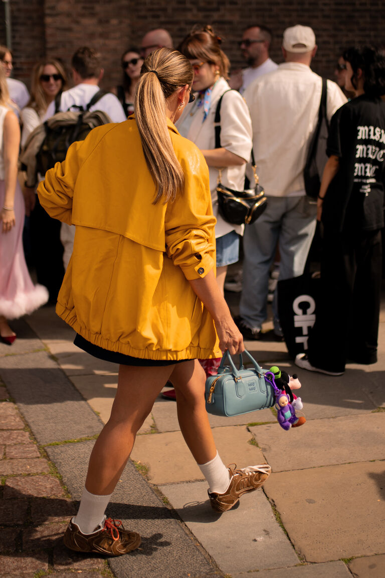 a woman at copenhagen fashion week wearing a yellow jacket, black skirt, and brown suede sneakers