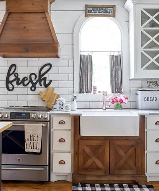 Farmhouse kitchen with wood and paint finished cabinets, arched window with curtain, wood hob hood, and large 'fresh' typo sign on backsplash.