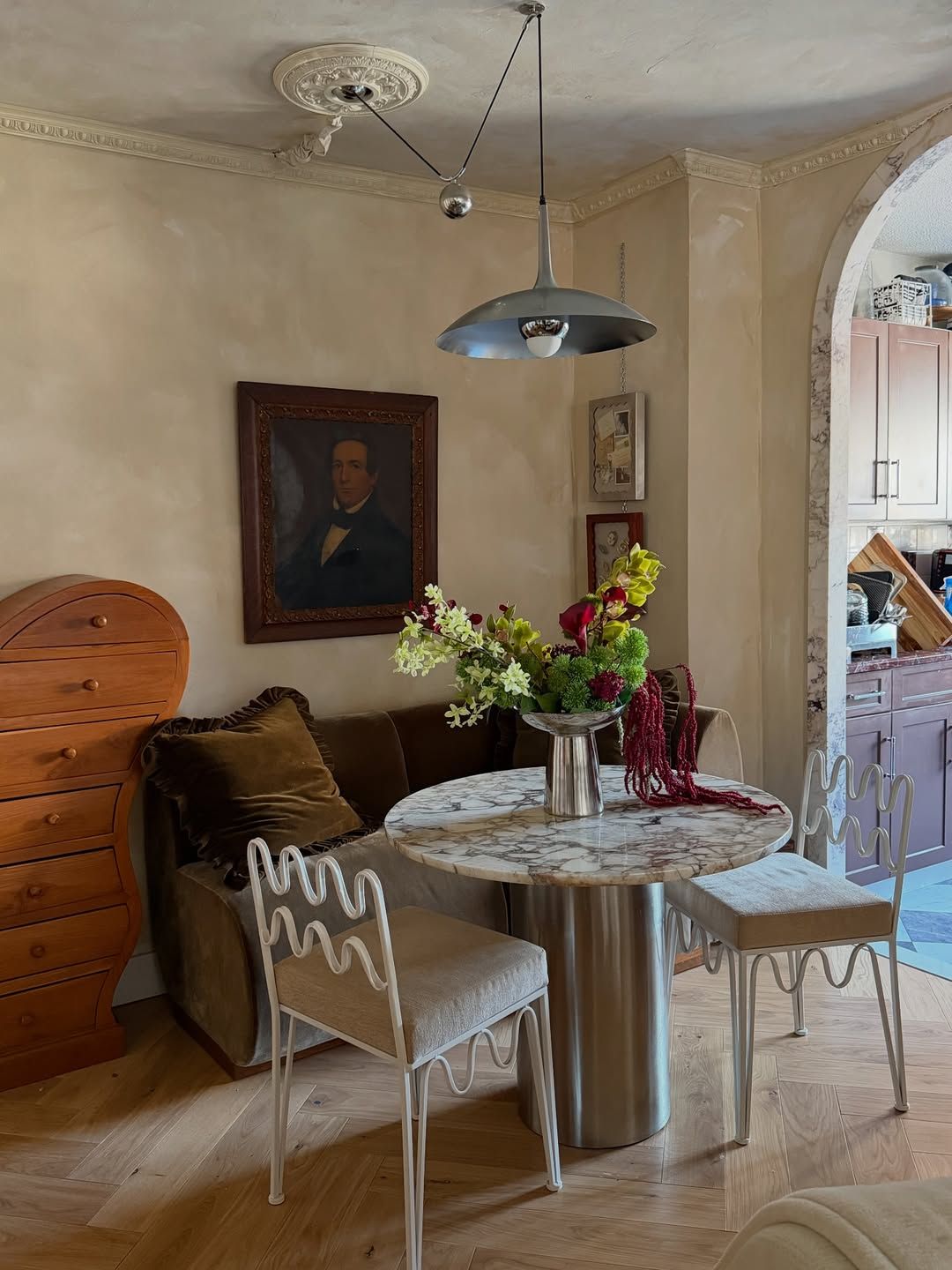 Image of a dining room with beige limewash walls, a marble circular table, wavy white dining chairs, and a velvet bench seat.