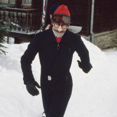 King Charles wearing a black ski suit walking through the snow with fake glasses, a fake mustache and comedy nose