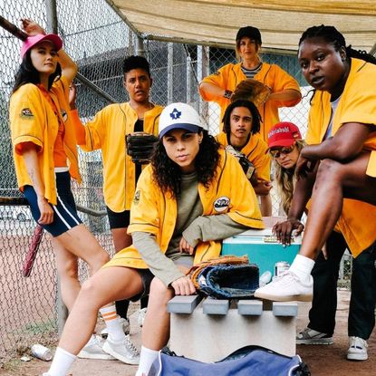A group of women wear yellow jerseys while posing in a baseball dugout, in the TV series 'Slo Pitch.'