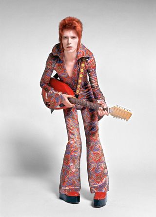 Portrait of David Bowie as playing the guitar wearing a bright patterned outfit.