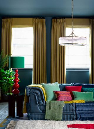 colorful living with room with blue walls and couch, turmeric colored drapes and blinds, pendant light, table lamp, bright cushions and throws, rug