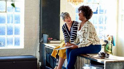 A couple of retired women laugh while sitting on a cabinet together listening to vinyl records.