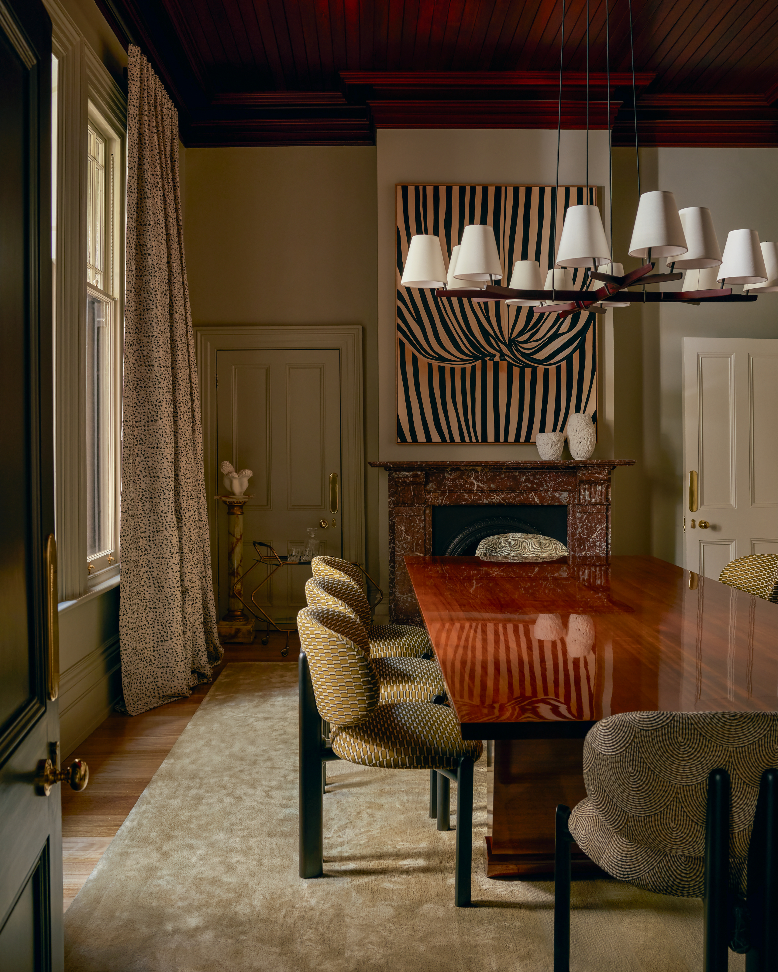 Dining room with beige walls, high gloss cherry wood table, black dining chairs upholstered with yellow patterned fabric, striped artwork and red marble fireplace