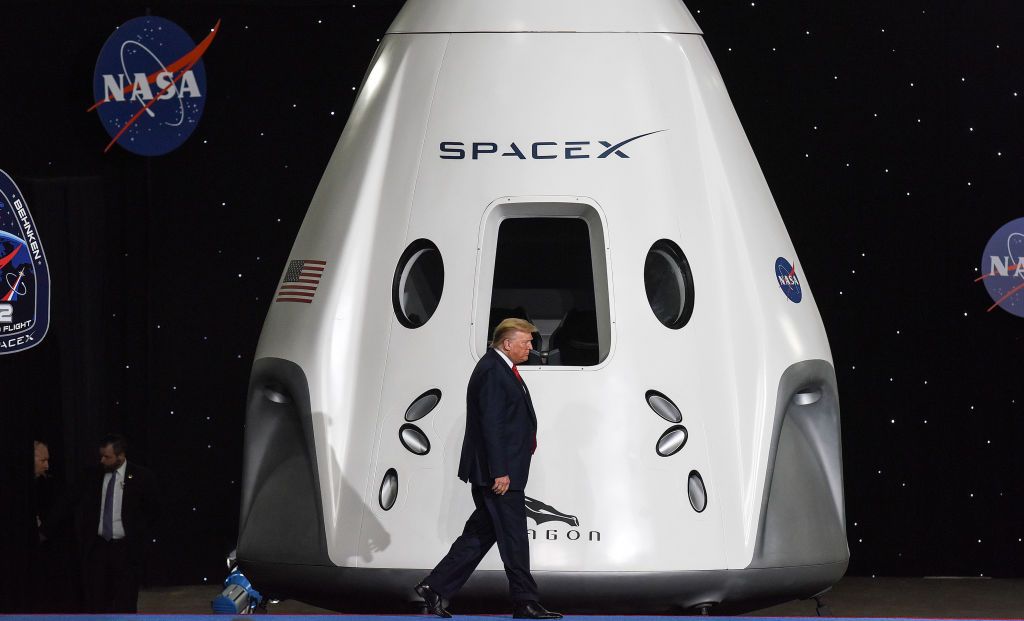 An orange man in a dark suit walks in front of a large white space capsule.