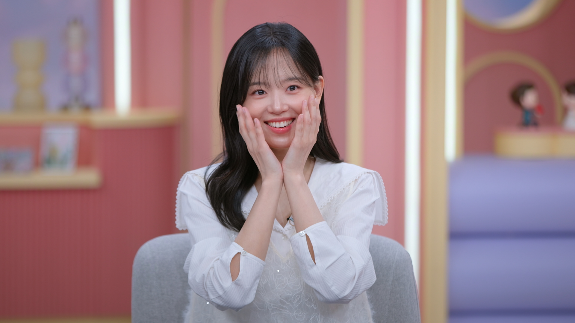 Co-host Kang Han-na holds her face in her hands while wearing a white button-down. She sits in a gray chair in a lounge with light-pink walls and lilac furniture, in 'Better Late Than Single.'
