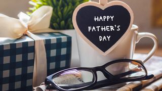 Photo of a gift box, glasses, and mug with a heart-shaped sign that reads Happy Father's Day