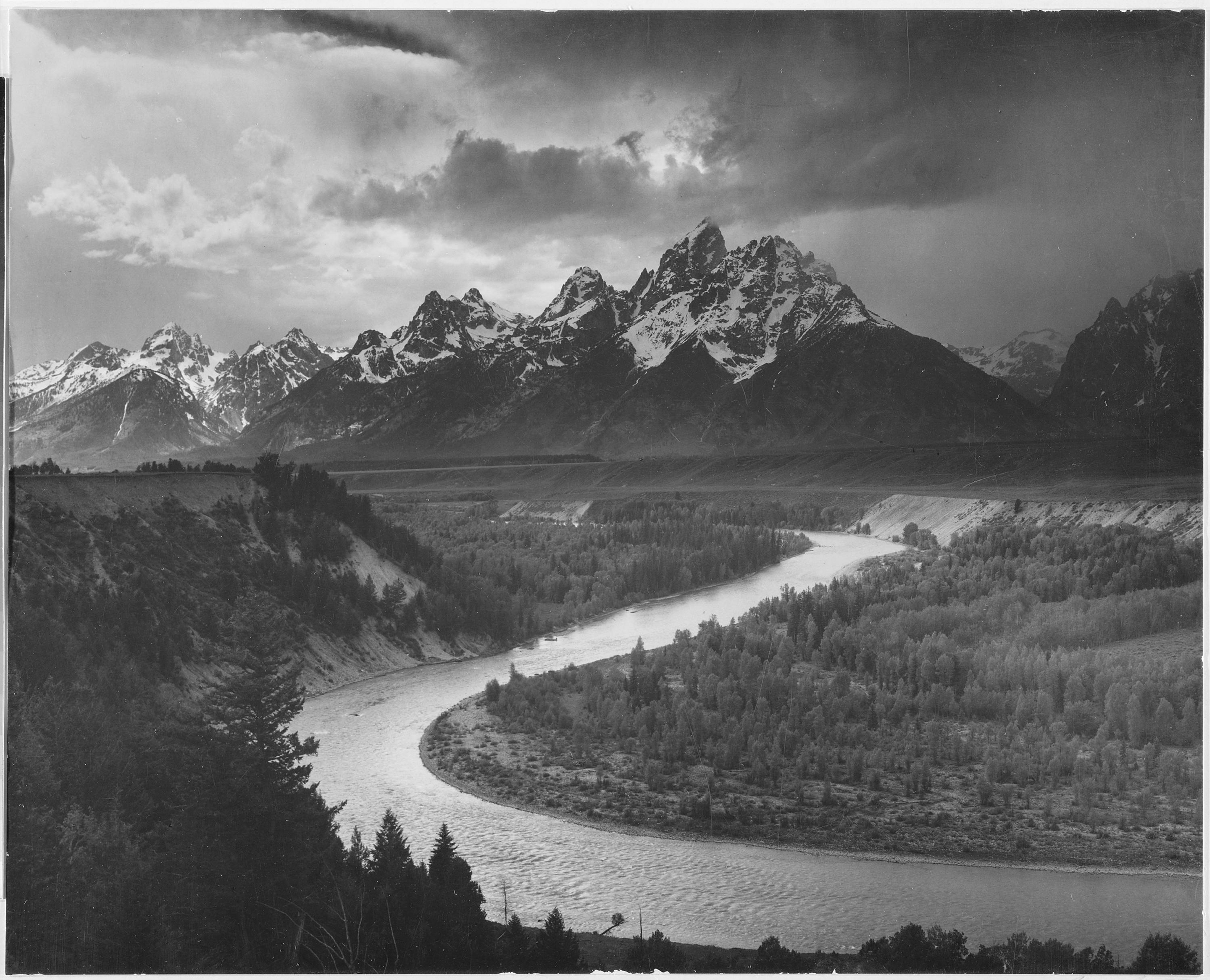 A black and white photograph of several of the peaks of the Teton Range covered in snow, the Snake River winds through the landscape and is surrounded by heavily forested lands