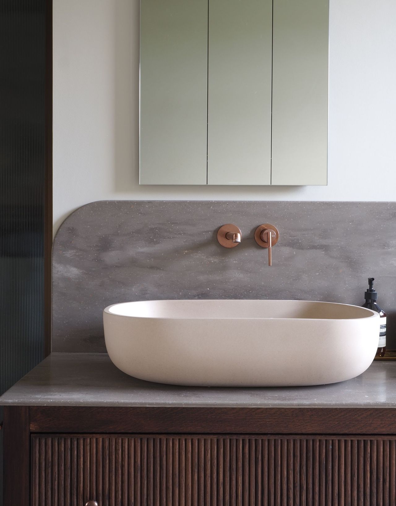 A modern bathroom sink on top of a vanity with wall-mounted bronze taps on a curved splashback underneath a mirrored cupboard