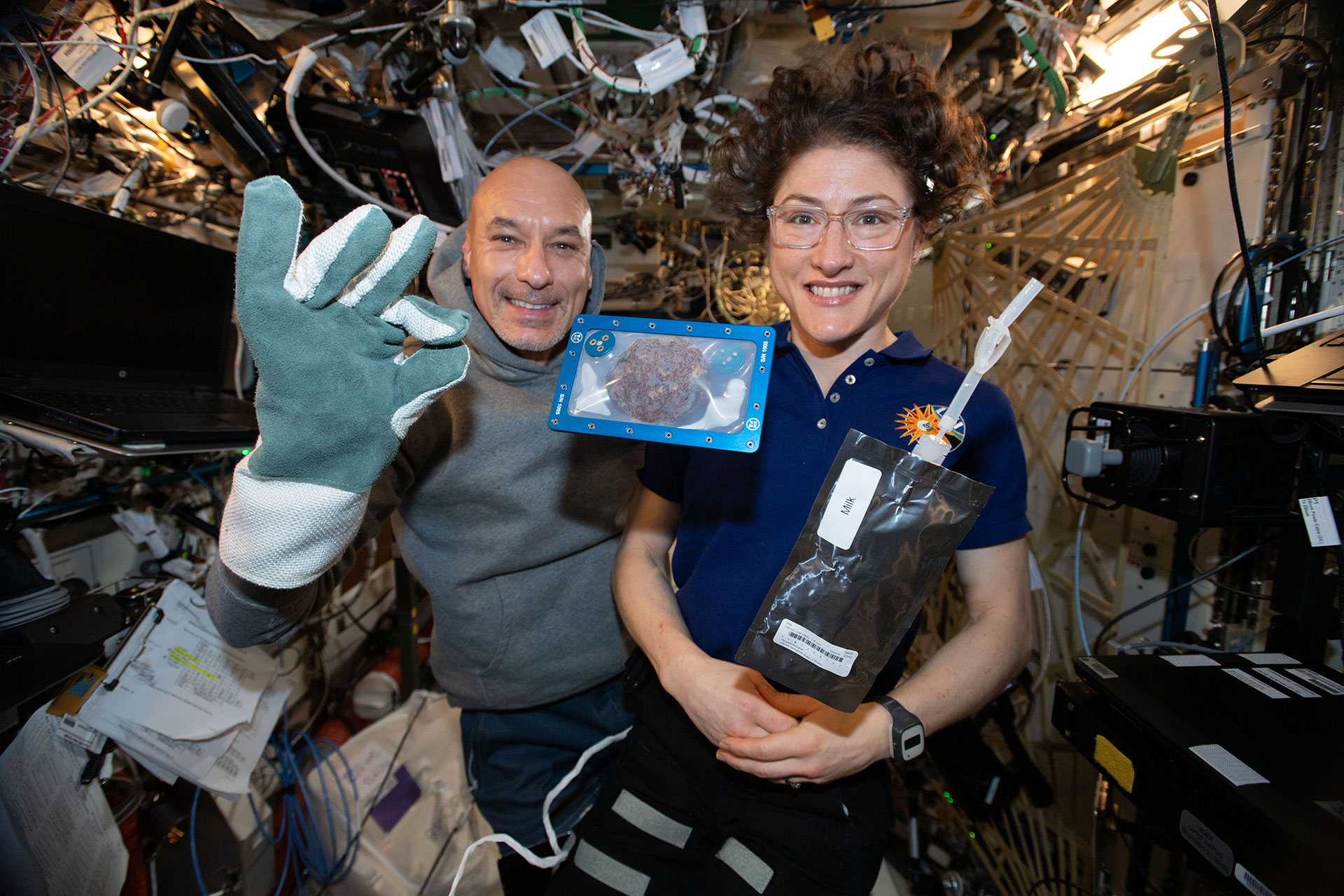 a man and a woman smile inside a space habitat, with a chocolate chip cookie enclosed in a case floating in front of them