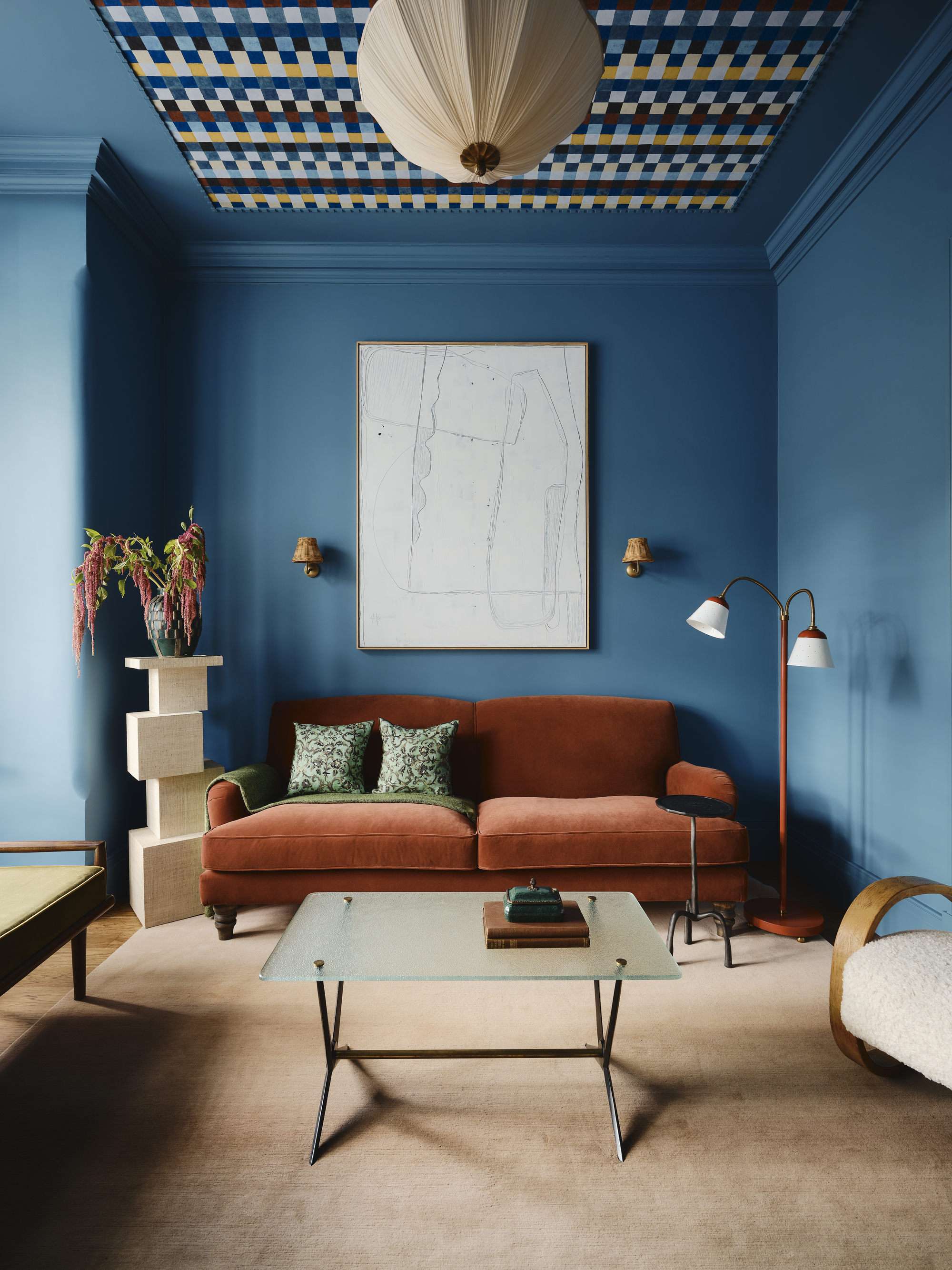 bold blue living room with patterned wallpaper on the ceiling, an orange velvet sofa with plinth holding flowers, floor lamp, glass and chrome coffee table, and tan colored carpet