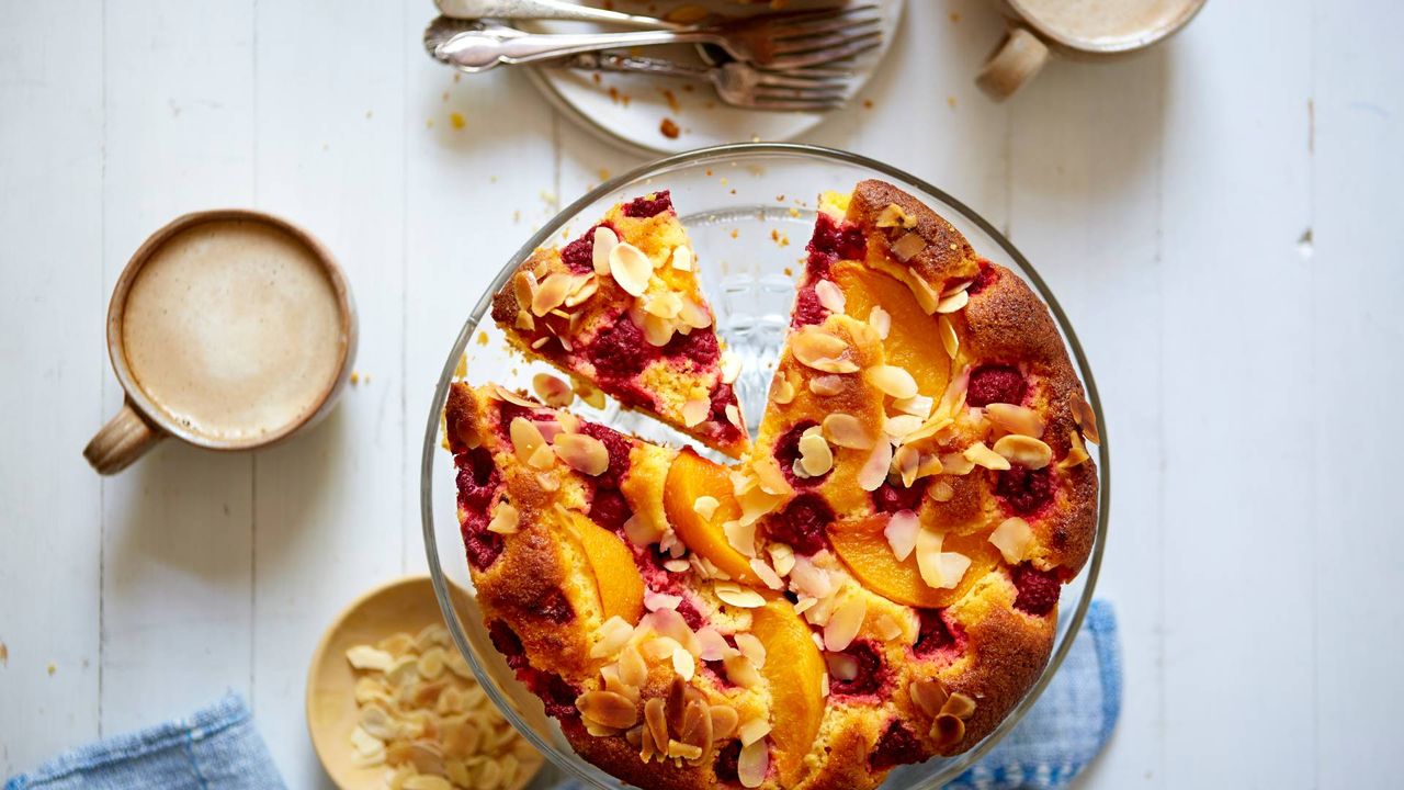 Raspberry polenta cake on a cake stand with two slices cut. Mug of coffee on the table and a small plate with flaked almonds.