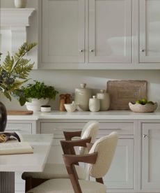 A white kitchen with cutting boards leaning against the backsplash, and an array of neutral-colored stone wear jars lined up around them. Two boucle barstool chairs with wooden arms in front, against a kitchen island. 