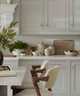 A white kitchen with cutting boards leaning against the backsplash, and an array of neutral-colored stone wear jars lined up around them. Two boucle barstool chairs with wooden arms in front, against a kitchen island.