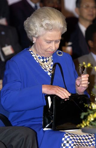 Queen Elizabeth wearing a blue coat and looking inside her black purse