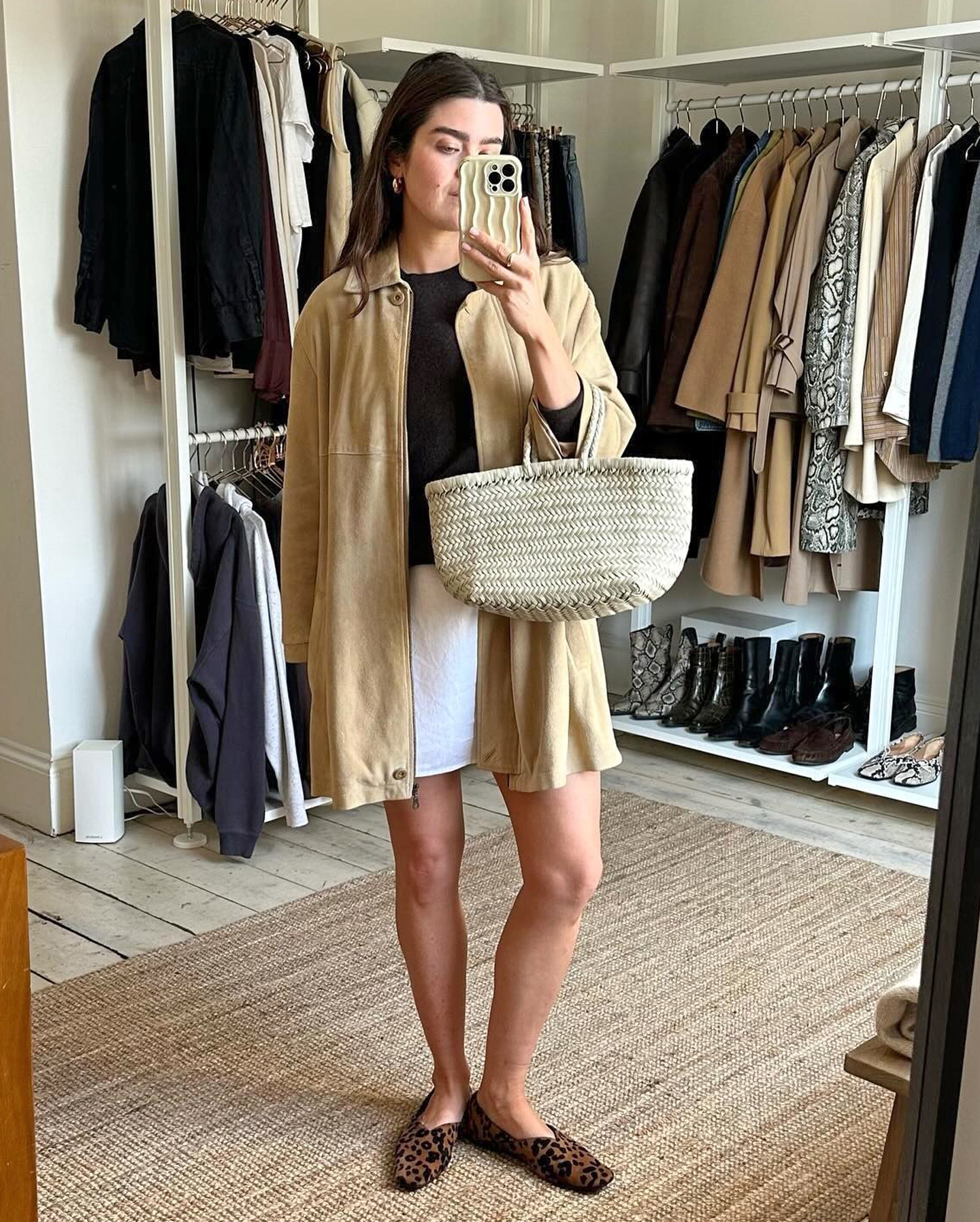 British style influencer Anna Newton poses for a mirror selfie in the closet of her London home wearing a tan suede jacket, black top, white woven tote bag, a white linen skirt, and leopard-print flats.