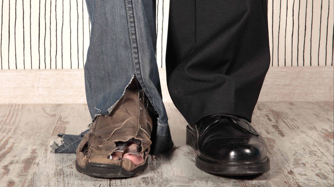 A mans feet seen from the knee down: One leg wears dress pants and polished work shoes, and the other torn jeans and sandals with holes in them.