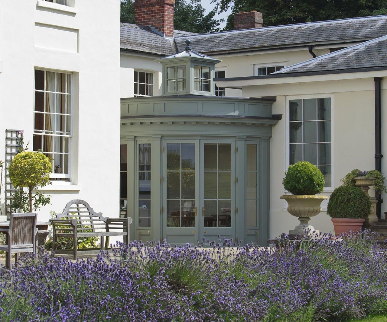 A curved sage green orangery with a glazed lantern cupola and brass-handled French doors sits between the wings of a white Georgian country house, fronted by sweeping lavender borders.