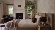 Image of a warm neutral bedroom with white walls that have mural wallpaper on it, a fireplace, and a dark brown chair in the corner, and a small tree.