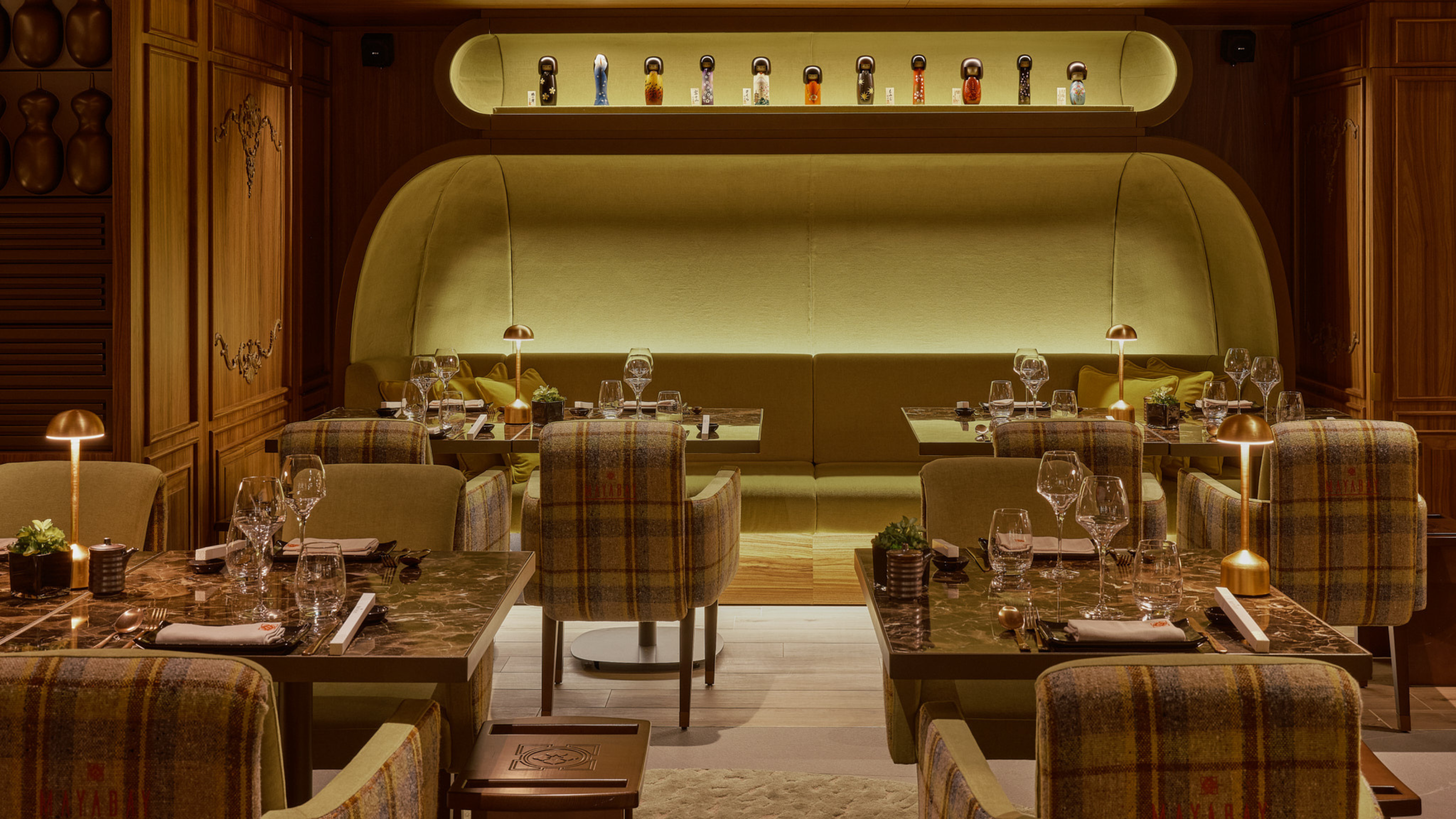 The MayaBay Courchevel restaurant with a space age meets traditional scheme with tartan armchairs at tables with table lamps and a large backlot green banquette seating area at the back with a backlot alcove above with decorative items