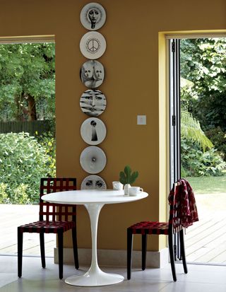 Yellow dining room with two open doors leading to a lush back yard. There is a small table with two red chairs inbetween the doors and a line of Fornasetti plates hanging above them.