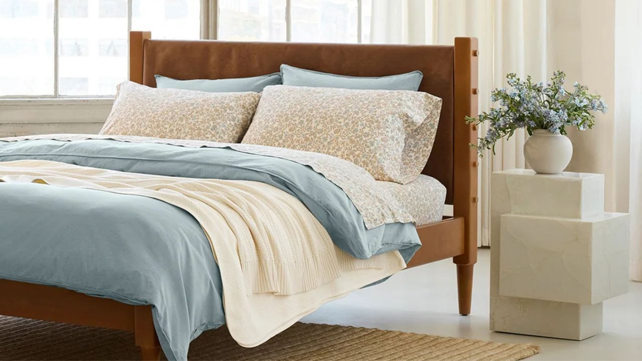 bed with blue and floral linen with sculptural stone side table with vase and foliage and curtains behind