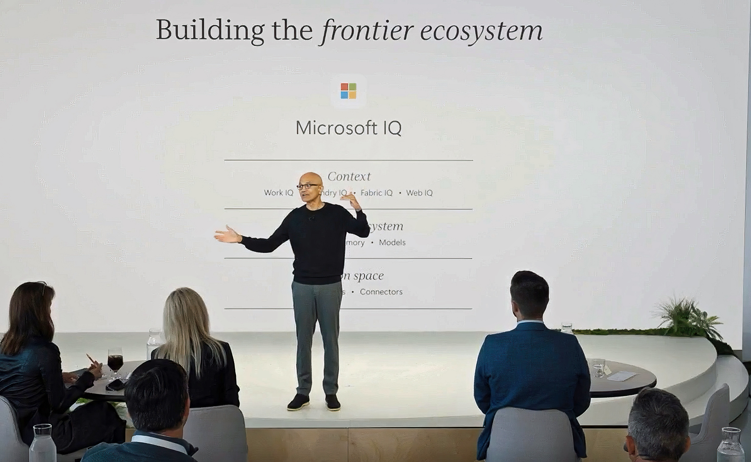 Satya Nadella presents on stage in front of a slide titled "Building the frontier ecosystem" displaying a "Microsoft IQ" diagram, facing a seated audience.