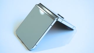 En grågrön Samsung Galaxy Z Flip 5 ligger halvt öppnad och nedåtvänd på ett bord.