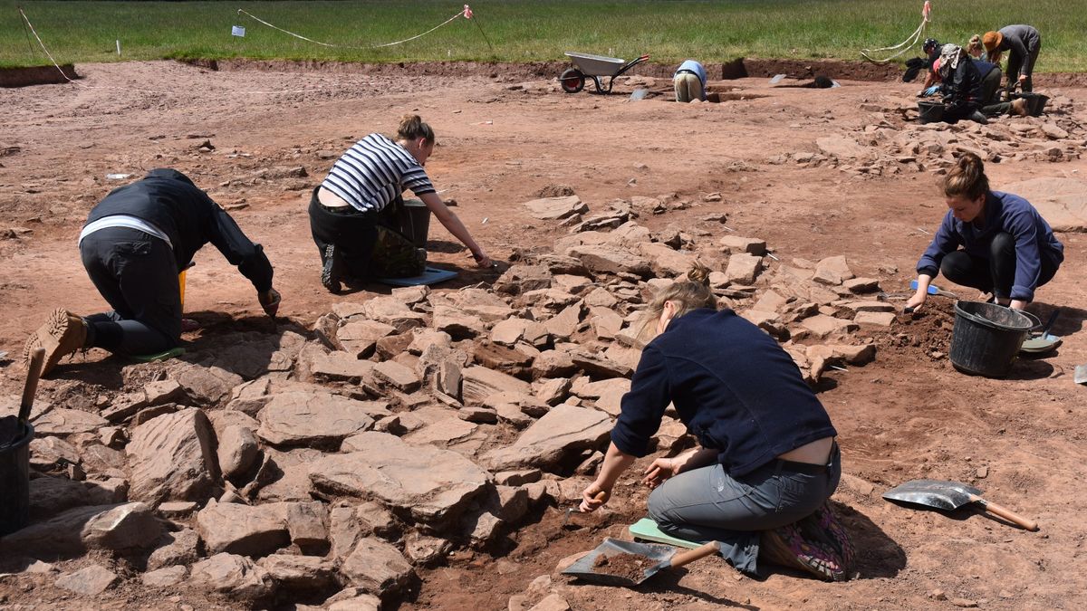 Close up of a Neolithic burial site at Dorstone Hill in the west of England. Here we see a large rectangle that has been dug up in a grassy field. There are a lot of people working on the site.