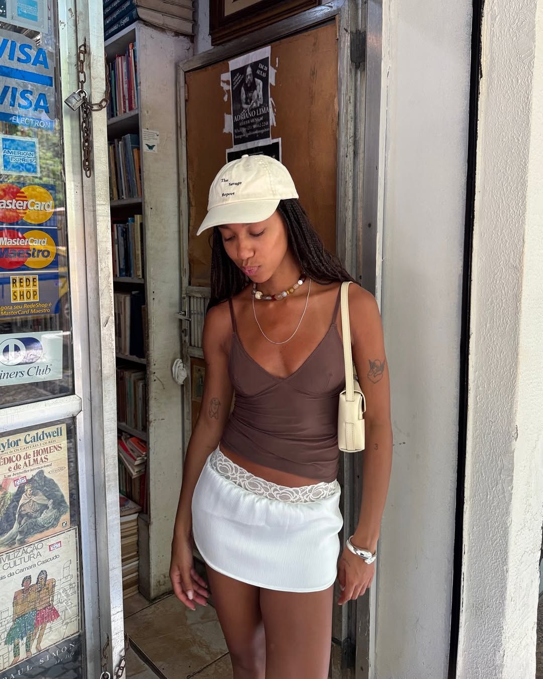Fashion person Amaka Hamelijnck is wearing a white satin mini skirt with lace trim, a brown V-neck top, a butter yellow cap and is carrying a butter yellow shoulder bag. Around her neck, she is wearing a beaded necklace and a silver bracelet around her wrist. She is standing in a doorway of an off-licence, looking down in a recent image on Instagram