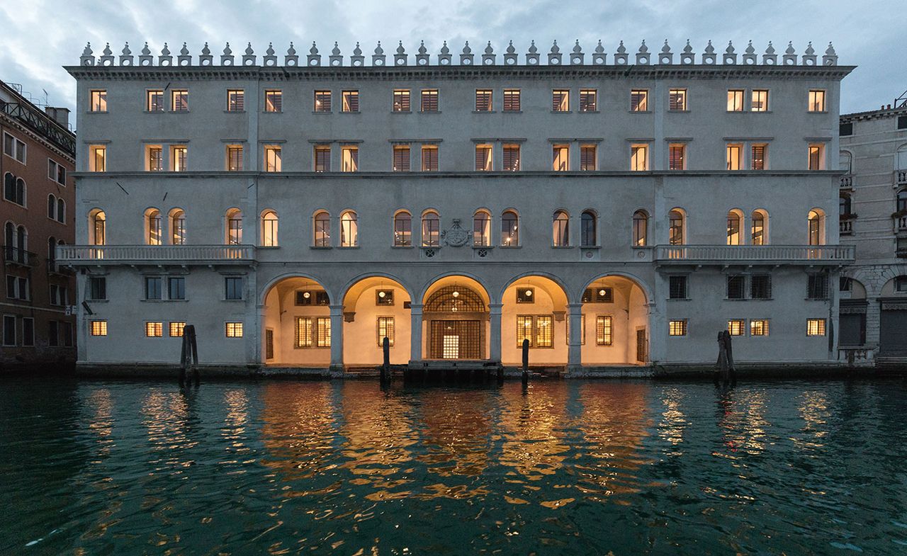 A 4-storey mansion-like building with Italian windows, balconies on the 1st floor with lights on in all the rooms. Photographed at dusk from the other side of the canal