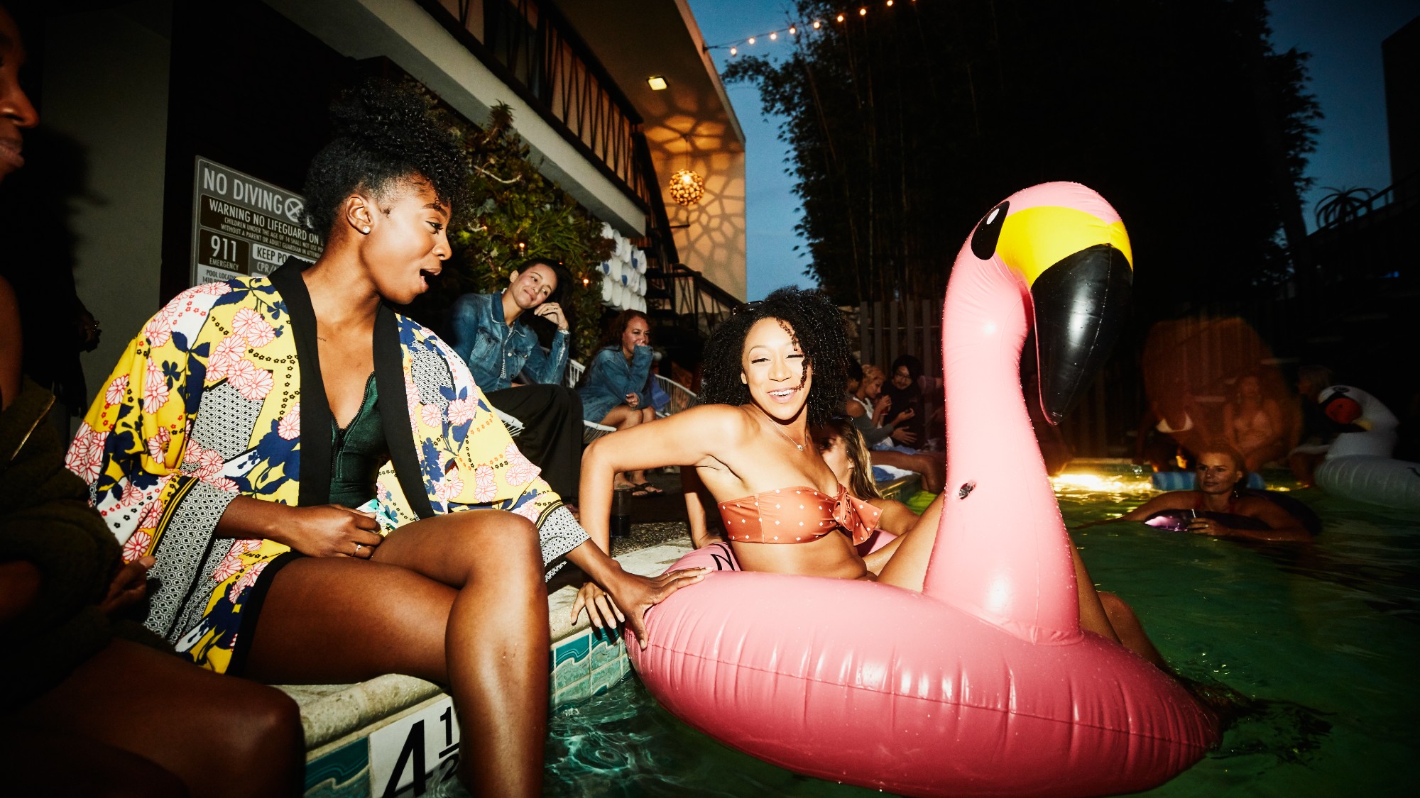 two woman having fun at a pool party. one of in a bikini inside of an inflatable flamingo in the pool. it is nighttime