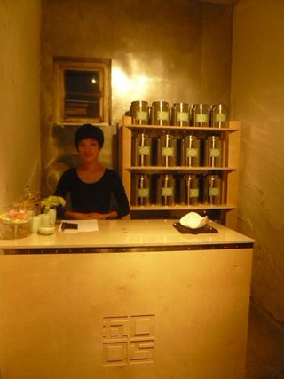 Woman standing behind a counter. Shelving unit in the background holding large silver cannisters