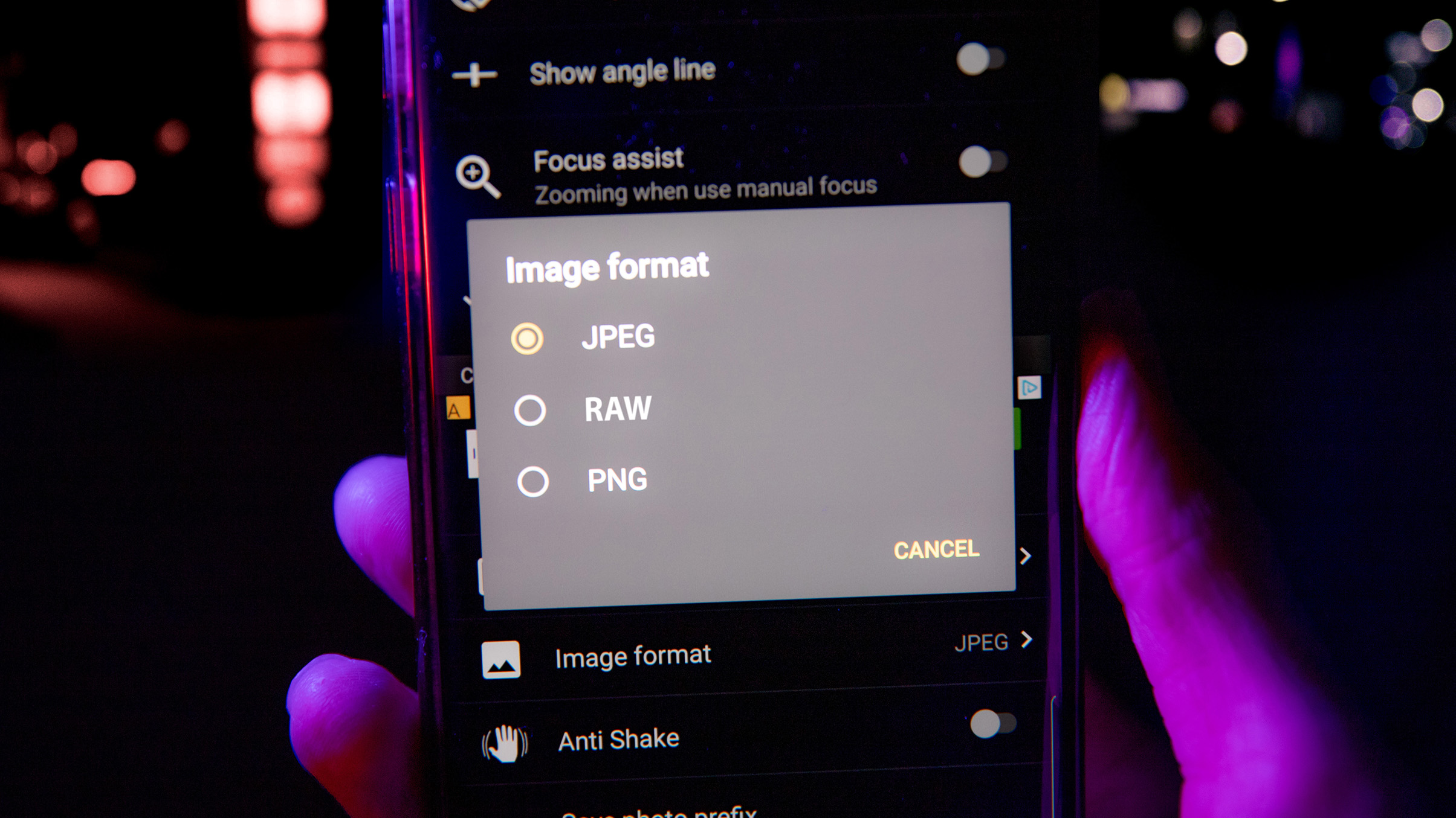 A hand holds a smartphone displaying an image format menu with options for JPEG, RAW, and PNG against a blurred background
